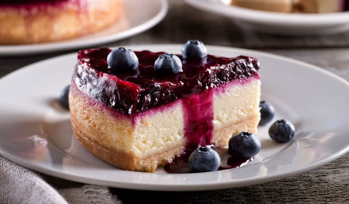A slice of homemade Blueberry Cheesecake on a white plate, featuring a golden-brown graham cracker crust, a thick and creamy layer of cheesecake filling, and a generous topping of fresh, plump blueberries. The cheesecake has a smooth, rich texture, and the blueberries provide a vibrant pop of color and a juicy, tangy flavor contrast