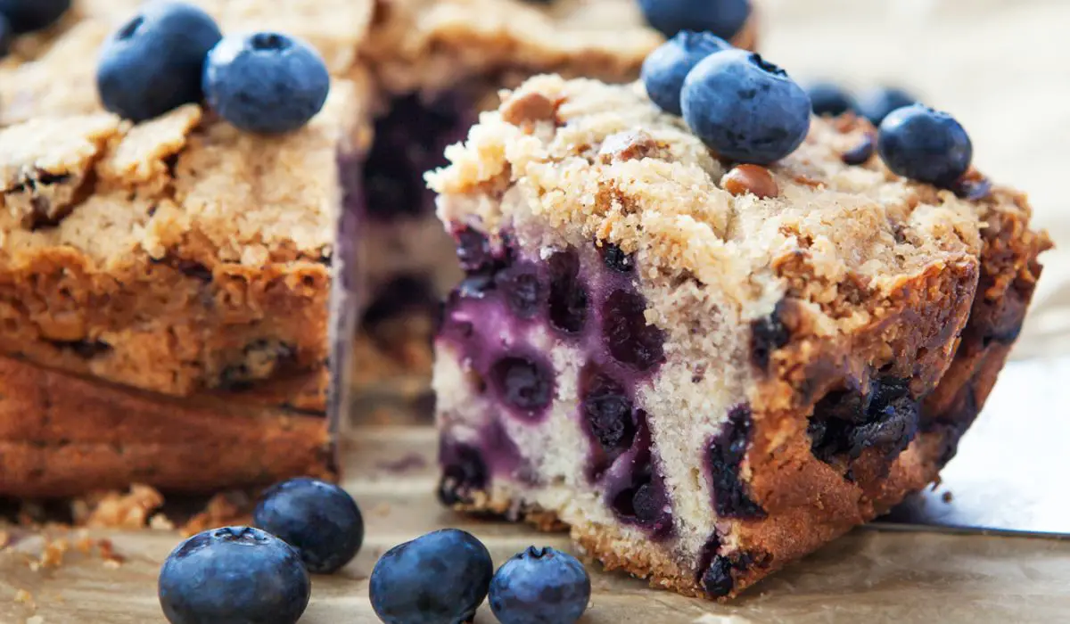 Image of a freshly baked Blueberry Coffee Cake on a white serving plate. The cake has a golden-brown streusel topping and is studded with juicy blueberries. Visible are the soft, moist texture of the cake and the rich, crumbly topping, creating a contrast of colors and textures. Slices of the cake reveal a tender, fluffy interior packed with blueberries, offering a visual promise of a delicious, sweet treat